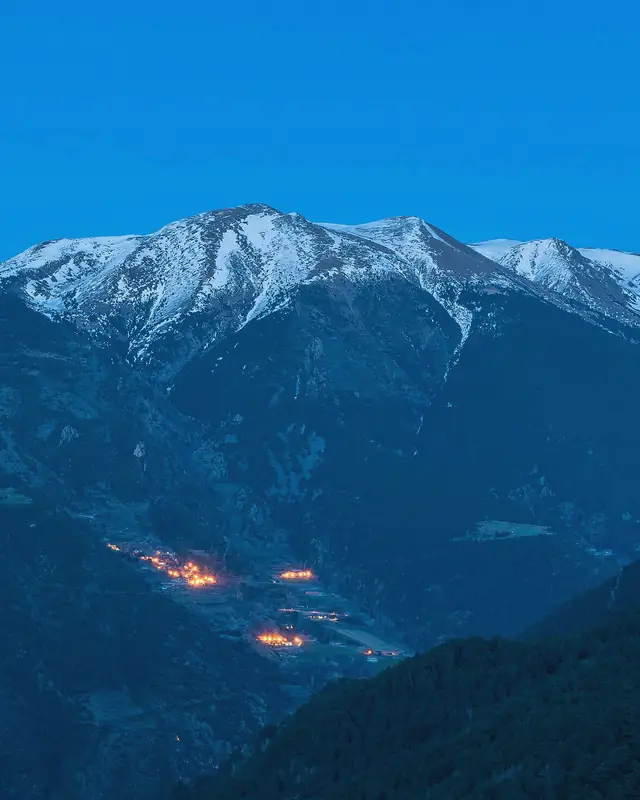 The snow-capped peaks of the Pyrenees in Andorra at the blue hour, the lights of a village at the bottom of the valley.