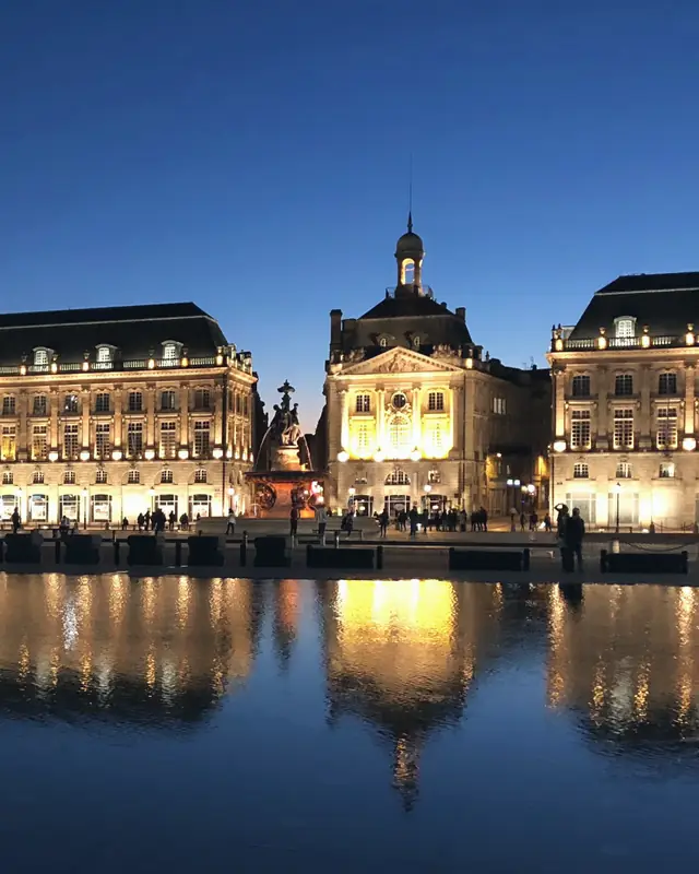 La place de la Bourse de Bordeaux à la nuit tombante : ses façades illuminées, la fontaine des Trois-Grâces et leur reflet dans l'eau.