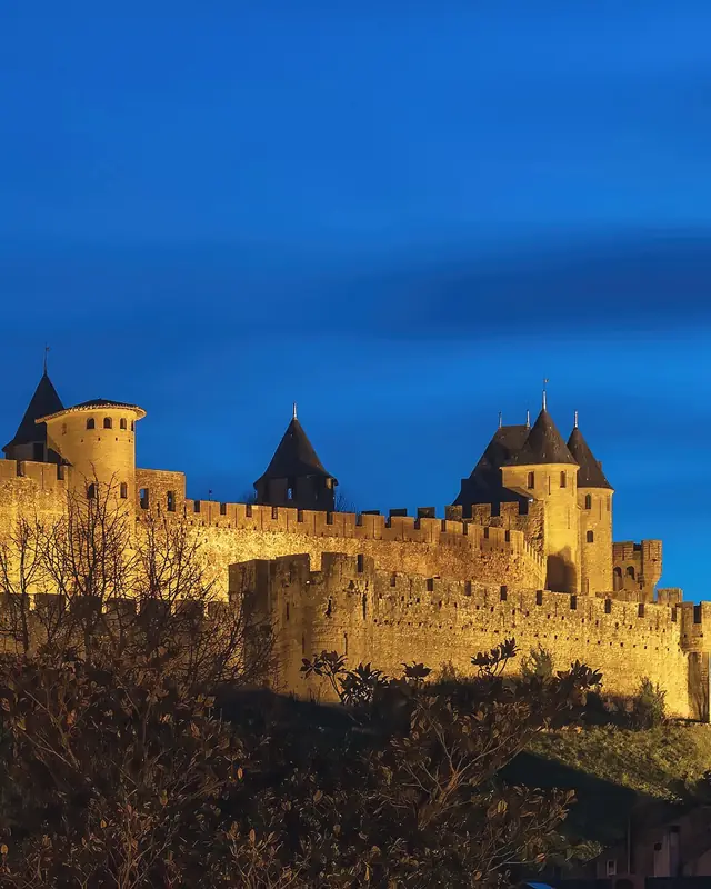 The Cité of Carcassonne lit up at the blue hour: its ramparts, its towers with pointed roofs and the Château Comtal under a deep blue evening sky.