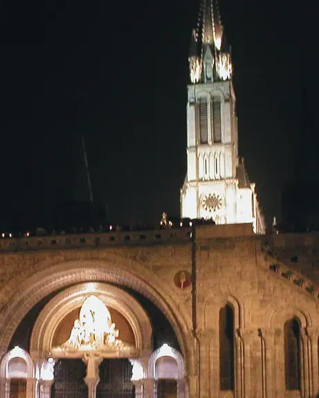 The Sanctuary of Lourdes at night: the Rosary Basilica and its great doorway, the lit spire of the Basilica of the Immaculate Conception above.