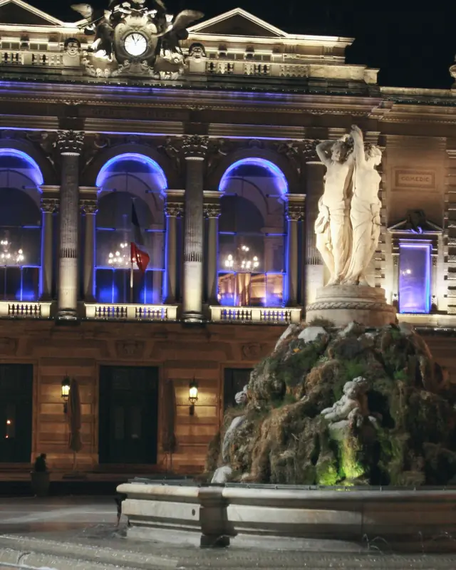 Place de la Comédie in Montpellier at night: the lit façade of the Opéra Comédie and the Three Graces fountain in front of it.