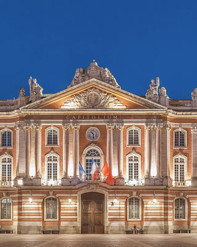 The façade of the Capitole in Toulouse lit up at the blue hour, its columns, its pediment and its flags.