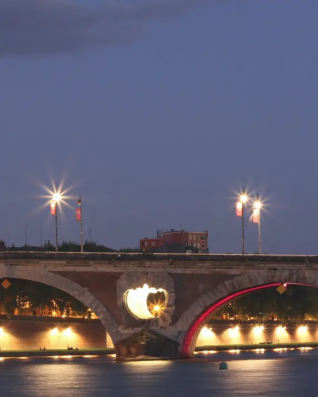 The Pont-Neuf in Toulouse at nightfall: its brick arches and lit round openings, the Garonne in the foreground.